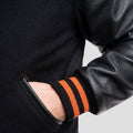 Classic black varsity jacket enhanced with black leather sleeves and bright orange ribbed accents, ideal for layering in any season
